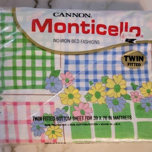 May include: A white twin-sized fitted bottom sheet with a green, blue, and pink gingham pattern. The sheet is made of 50% polyester and 50% cotton and is made in the USA. The packaging says "CANNON Monticello NO-IRON BED FASHIONS".