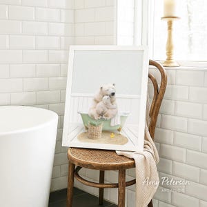 A framed image of a polar bear sitting in a bathtub, with a basket of toys nearby, is displayed on a wooden chair in a bathroom setting.