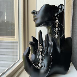 May include: A pair of silver-toned dangle earrings featuring a large circular hoop frame. Each earring includes two smaller circular rings connected by silver links, with a small, textured, spherical bead centered within each of the smaller rings.