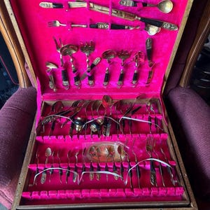 May include: An open wooden cutlery case with a bright pink velvet interior. The case contains an assortment of gold-toned silverware, including forks, spoons, and knives. The handles are black and gold. The case is sitting on a maroon chair.