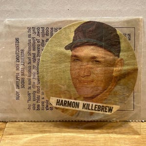 Pode incluir: Um cartão de beisebol vintage com Harmon Killebrew, em uma manga protetora de plástico. O cartão apresenta uma imagem circular de Killebrew usando um boné. O nome "HARMON KILLEBREW" é impresso em uma etiqueta branca.
