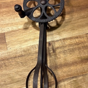 Puede incluir: Batidora de huevos antigua de hierro fundido con un engranaje circular y un mango. La batidora tiene un aspecto oscuro y envejecido, lo que sugiere que es vintage. El diseño incluye un mango para girar y un mecanismo similar a un batidor.
