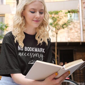 Può includere: Una maglietta nera con la scritta "No Bookmark Necessary" e una grafica di un libro. La persona indossa una gonna azzurra e legge un libro con pagine bianche. Lo sfondo include un'ambientazione da caffè con tavoli e sedie in legno.