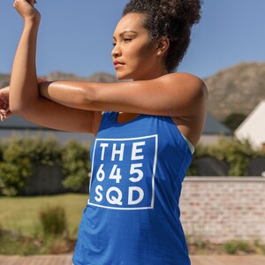May include: A blue tank top with the text "THE 645 SQD" in a white square. The tank top is being worn by a person stretching outdoors. The background includes a pool, mountains, and a clear blue sky.