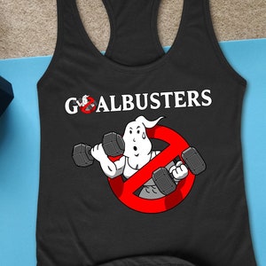 May include: Black racerback tank top with the text "GOALBUSTERS" and a cartoon ghost lifting dumbbells inside a red no-entry sign. Two black dumbbells and a blue yoga mat are also visible.