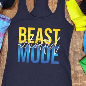May include: A navy blue racerback vest with the text "BEAST ACTIVATED MODE" in yellow, white, and blue. The image includes a pair of 6 kg dumbbells, colourful resistance bands, and patterned workout shorts.