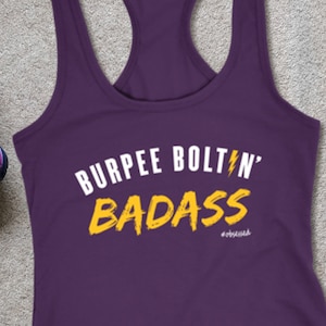 May include: A purple racerback tank top with the text "BURPEE BOLTIN' BADASS" in white and yellow. The image also includes purple athletic shoes with blue laces, a blue yoga mat, and two teal dumbbells.