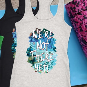 May include: Three racerback tank tops, one black and two gray, with the text "WE'RE NOT THERE YET!" on the front. The text is over a colorful abstract design. Also shown are exercise bands and a pink camo foam roller.