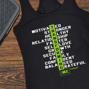 May include: Black racerback tank top with white text listing motivational words, with the word "STRONG" highlighted in green. A black exercise step is on the left side of the image, set against a wood grain background.
