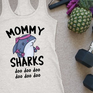 May include: A light gray tank top with the words "MOMMY SHARKS" and a cartoon shark graphic. The shark is wearing a pink headband and holding dumbbells. Other items include two 9 kg dumbbells, a pineapple, and a jump rope.