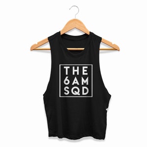 Pode incluir: Top curto preto sem mangas com o texto "THE 6 AM SQD" em um quadrado branco. A parte superior está pendurada em um cabide de madeira contra um fundo branco. O texto está em negrito, fonte sem serifa.