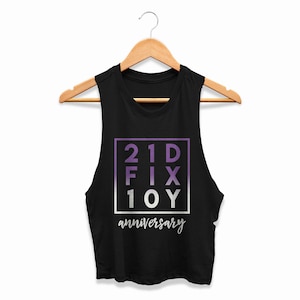 May include: Black sleeveless crop top with a wooden hanger. The top features a purple and white graphic that reads "21D FIX 10Y anniversary". The text is inside a purple and white square.