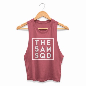 Pode incluir: Um top curto sem mangas, rosa empoeirado, com o texto "THE 5 AM SQD" num quadrado branco. O top está pendurado num cabide de madeira sobre um fundo branco. O tecido parece ser um material macio e leve.