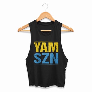 May include: Black sleeveless vest top with the text "YAM SZN" in yellow and blue. The top is hanging on a wooden hanger against a white background. The text has a distressed, vintage appearance.