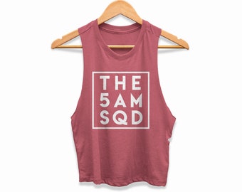 May include: A sleeveless, dusty rose crop top with the text "THE 5 AM SQD" in a white square. The top is hanging on a wooden hanger against a white background. The fabric appears to be a soft, lightweight material.