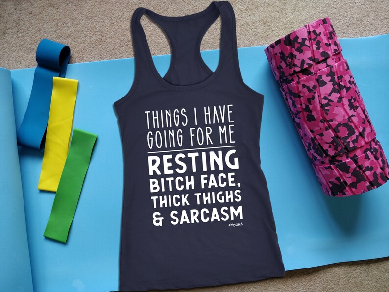 May include: Navy blue racerback tank top with white text: "THINGS I HAVE GOING FOR ME RESTING BITCH FACE, THICK THIGHS & SARCASM." Also shown are blue, yellow, and green resistance bands, and a pink camo foam roller.