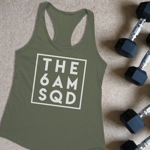 May include: An olive green racerback vest with the text "THE 6AM SQD" in a white square. Two pairs of black dumbbells with silver handles are next to the vest. The vest is made of a soft material.