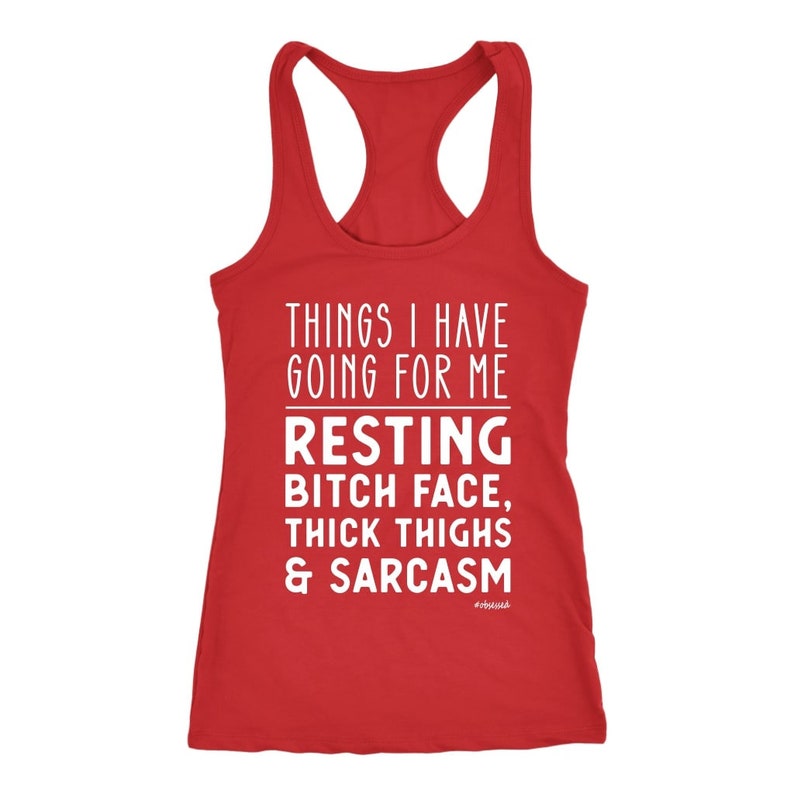 May include: A red racerback tank top with white text that reads: "THINGS I HAVE GOING FOR ME RESTING BITCH FACE, THICK THIGHS & SARCASM." The tank top has a scoop neck and a racerback design.