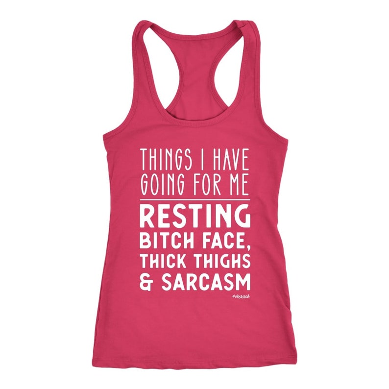 May include: A raspberry-colored racerback tank top with white text. The text reads "THINGS I HAVE GOING FOR ME" above "RESTING BITCH FACE, THICK THIGHS & SARCASM". The tank top has a scoop neck and a racerback design.