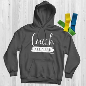 Puede incluir: Sudadera con capucha gris oscuro con las palabras "Coach ALL YEAR" en escritura blanca. La sudadera tiene un bolsillo delantero y una capucha con cordón. Un conjunto de bandas de resistencia coloridas en amarillo, verde y azul está al lado de la sudadera.