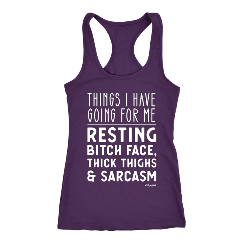 May include: A purple racerback tank top with white text. The text reads "THINGS I HAVE GOING FOR ME" above "RESTING BITCH FACE, THICK THIGHS & SARCASM". The word "#obsessed" is at the bottom.