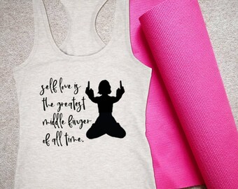 May include: A light gray racerback tank top with the text "self love is the greatest middle finger of all time" and a black silhouette of a person. A rolled-up pink yoga mat is next to the tank top.