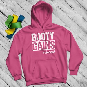 Puede incluir: Una sudadera con capucha rosa con el texto blanco "BOOTY GAINS" y "#obsessed". La sudadera tiene un bolsillo delantero y una capucha con cordón. Bandas de resistencia amarillas, verdes y azules en el fondo.