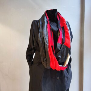 Peut inclure: Un manteau noir avec une écharpe rouge, noire et grise drapée autour du cou. L'écharpe présente un motif abstrait audacieux avec un mélange de textures et de couleurs. Le manteau est boutonné et possède une ceinture.