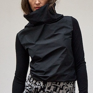 Op de afbeelding: Een zwart shirt met lange mouwen, een hoge kraag en duimgaten in de boorden, gecombineerd met een zwarte rok met een wit geometrisch blokpatroon dat vervaagt naar een effen zwarte zoom. Het ensemble bestaat uit een aansluitend lijfje en een rechte rok.