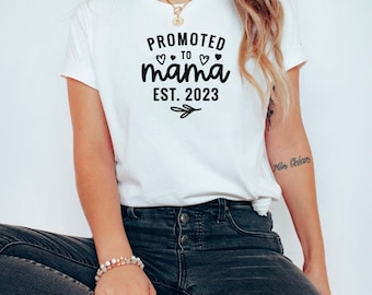 May include: White t-shirt with black text that reads "Promoted to mama est. 2023" with three small hearts above the word "mama".