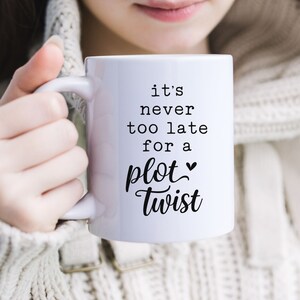 May include: White ceramic mug with a black inscription that reads "it's never too late for a plot twist" with a heart symbol. The mug is being held by a person wearing a white sweater.