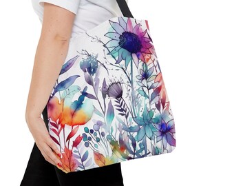 May include: A white tote bag featuring a vibrant watercolor floral design. The artwork showcases a variety of colorful flowers and foliage in shades of blue, purple, orange, and red. The bag has a black strap for carrying.