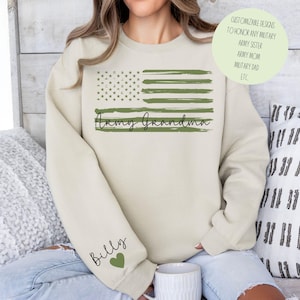 May include: A beige sweatshirt with a green and white American flag design. The text "Army Grandma" is printed below the flag. The left sleeve has the text "Billy" and a green heart.