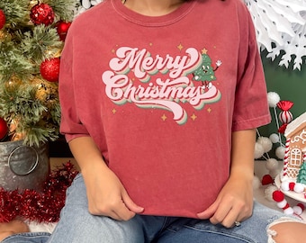 May include: A red t-shirt with a retro-style "Merry Christmas" graphic featuring a cartoon Christmas tree with a smiling face and waving arms.