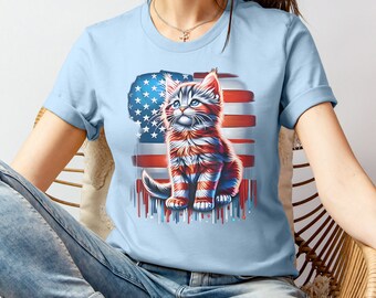 May include: A light blue t-shirt with a graphic of a kitten sitting in front of an American flag. The kitten is red, white, and blue, and the flag is dripping with paint.