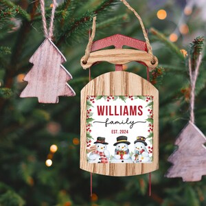 May include: A wooden Christmas ornament shaped like a sled with a red and white design. The ornament features a personalized message that reads "Williams family est. 2024" and a graphic of three snowmen.