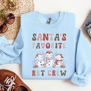 May include: Light blue sweatshirt with a Christmas theme. It features three snowmen with red hats and the text "Santa's Favorite RBT Crew".