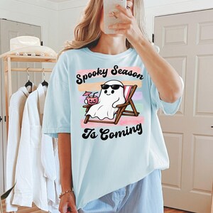 May include: A light blue t-shirt featuring a graphic of a white ghost wearing black sunglasses, relaxing in a wooden beach chair while holding a pink jack-o'-lantern drink with an umbrella. The text reads Spooky Season Is Coming.