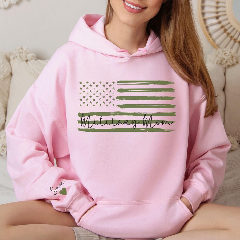 May include: Pink hooded sweatshirt with a green and white American flag design and the text "Military Mom". The sleeve has the name "Sam" and a green heart.