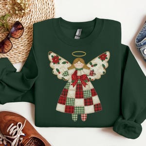 May include: A dark green sweatshirt featuring a patchwork angel design. The angel has a gold halo, blonde hair, and a red bow. The wings are decorated with poinsettias and the dress is a patchwork of red, green, and white squares.