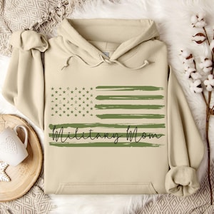 May include: A beige hoodie with a green and white American flag design and the text "Military Mom" in black script.