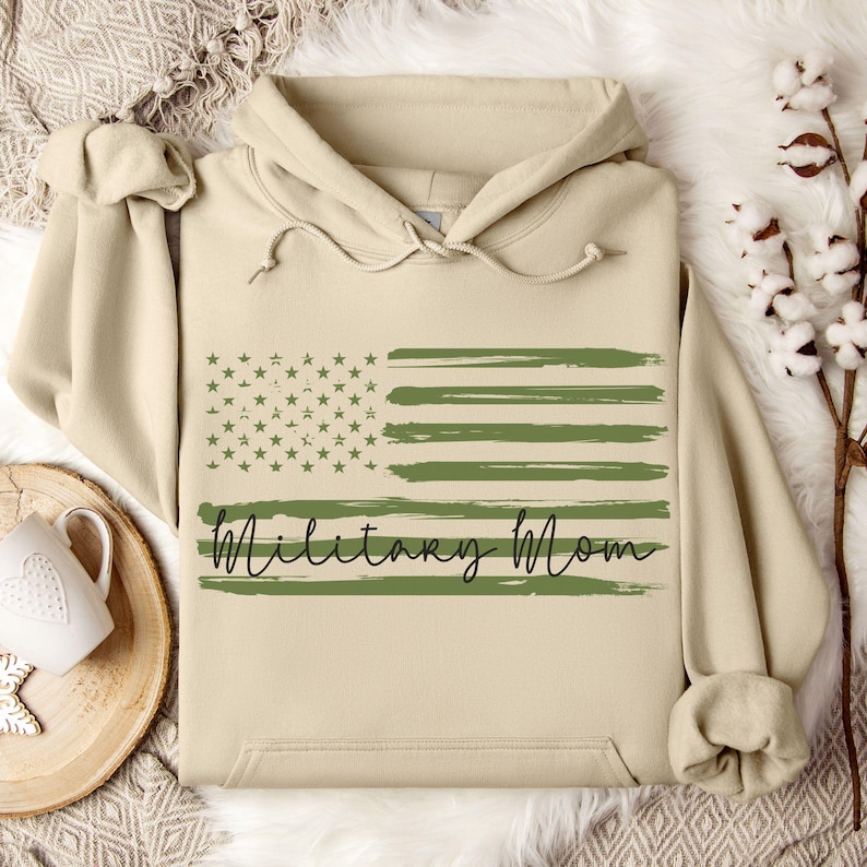 May include: A beige hoodie with a green and white American flag design and the text "Military Mom" in black script.