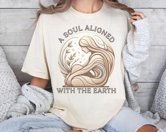 May include: A beige t-shirt with a graphic design of a woman sitting with her hair flowing around her. The text "A Soul Aligned With The Earth" is printed on the shirt.