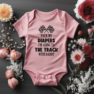 Puede incluir: Un body rosa para bebé con texto negro que dice "Pack my diapers I'm going to the track with daddy". El body tiene un gráfico de dos banderas de carreras a cuadros.