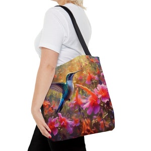 May include: A tote bag with a colorful hummingbird in flight over a field of pink and orange flowers. The bag has black straps.