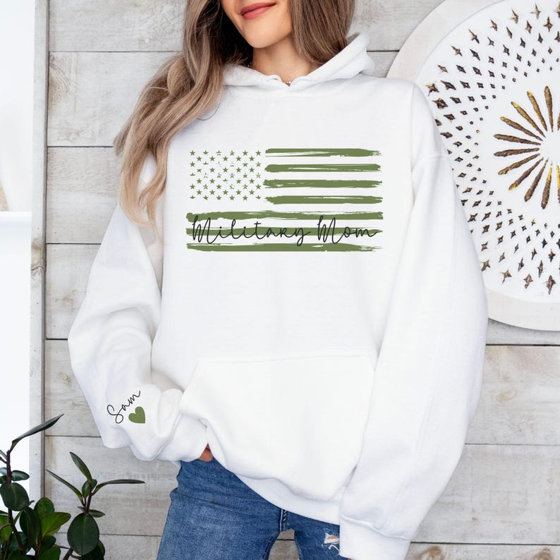May include: White hooded sweatshirt with a green and white American flag design and the text "Military Mom". The sleeve has a green heart with the text "Sam" on it.