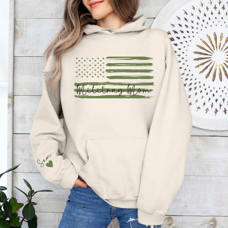 May include: A cream-colored hoodie with a green and white American flag design and the text "Military Mom". The hoodie has a pocket on the front and a green heart with the name "Sam" on the left sleeve.