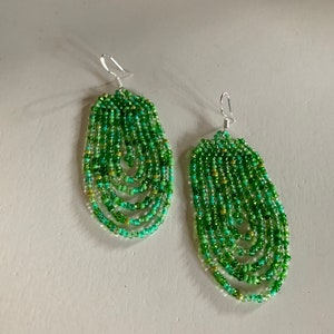 May include: A pair of green beaded earrings with a teardrop shape. The earrings are made of small, round beads in shades of green, with a silver hook.