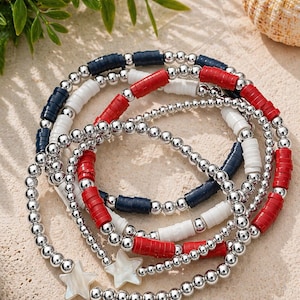 May include: A set of four beaded bracelets in red, white, and blue, with silver-colored beads. Two bracelets feature star-shaped charms. The bracelets are arranged on a light-colored surface, with greenery and a seashell in the background.