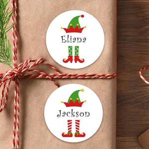 May include: Two white circular stickers with elf-themed designs are displayed on a wrapped gift. Each sticker features an elf hat, name, and elf legs. The names "Eliana" and "Jackson" are printed in black. The gift is wrapped in brown paper and tied with red and white twine.